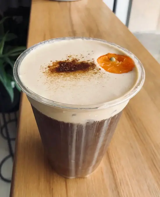 Close up of a cold foam topped cold brew in a plastic cup with spices sprinkled on top and a floating piece of orange fruit.