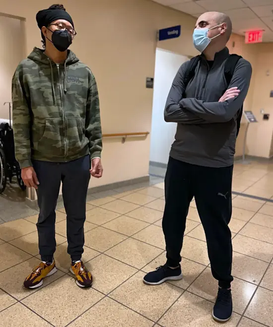 Lem wears a camouglage hoodie and face mask in a hospital hallway with Nathanael, in a pullover and mask with backpack.