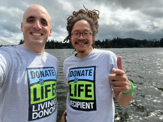 Nathanael wears a shirt saying donate life: living donor. Lem wears a shirt saying donate life: recipient. They stand by a lake.