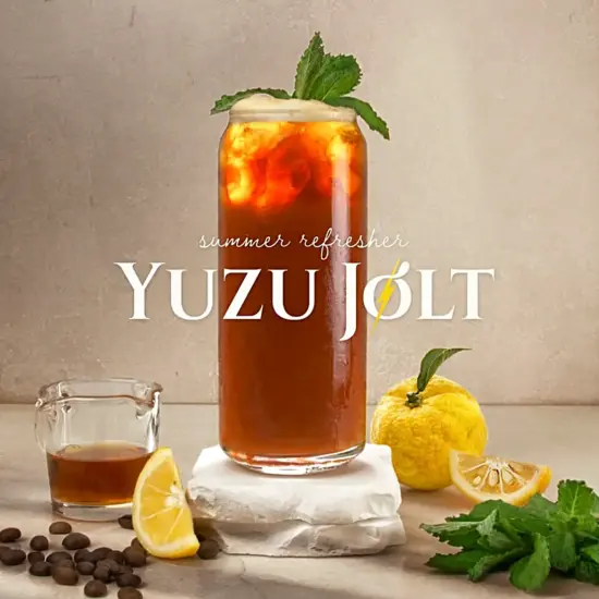 An iced drink in a tall can-shaped glass with mint sprig. Around the drink yuzu fruits and mints leaves are arranged.