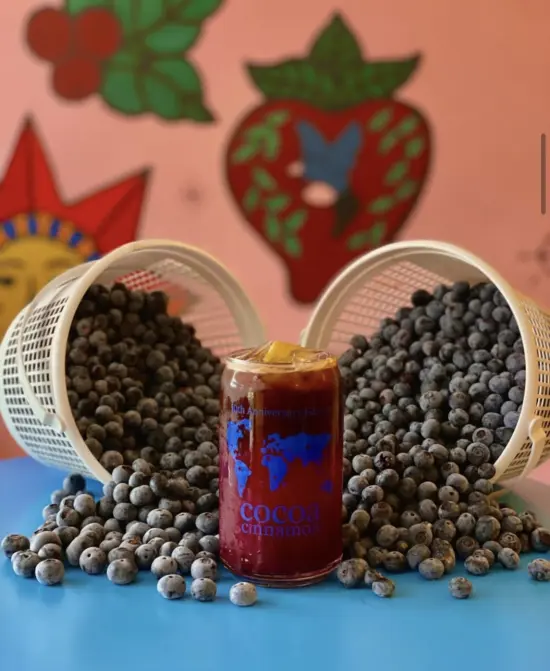 A short can glass with a dark iced drink is surrounded by blueberries spilling from two baskets.
