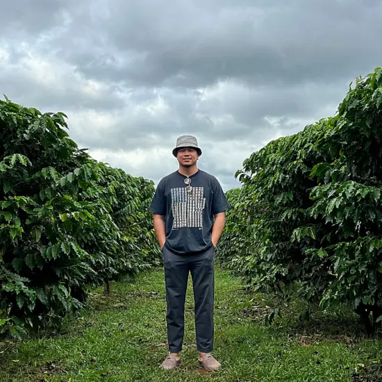 Emil Vanta in casual clothes and a bucket hat stands between coffee trees. A cloudy sky looms behind.