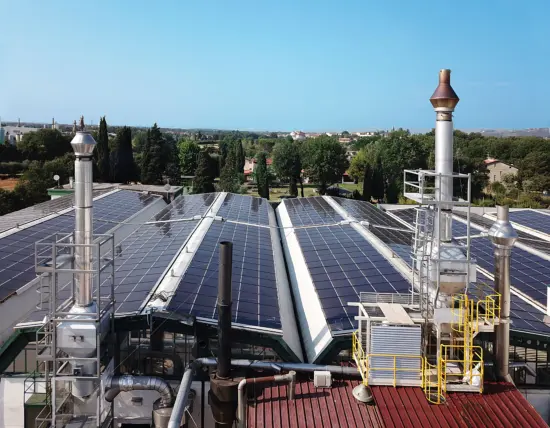 A view of industrial equipment with smokestack type structures and with solar panels splayed out behind on a rooftop.