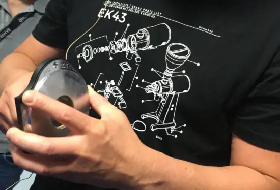 A person holds grinder parts. There is a Mahlkonig grinder labeled on his tshirt.