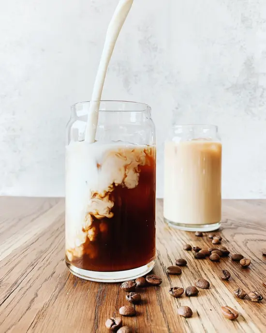 Milk being poured into a glass container of cold coffee on a wooden surface with scattered beans and another glass of milk behind it.