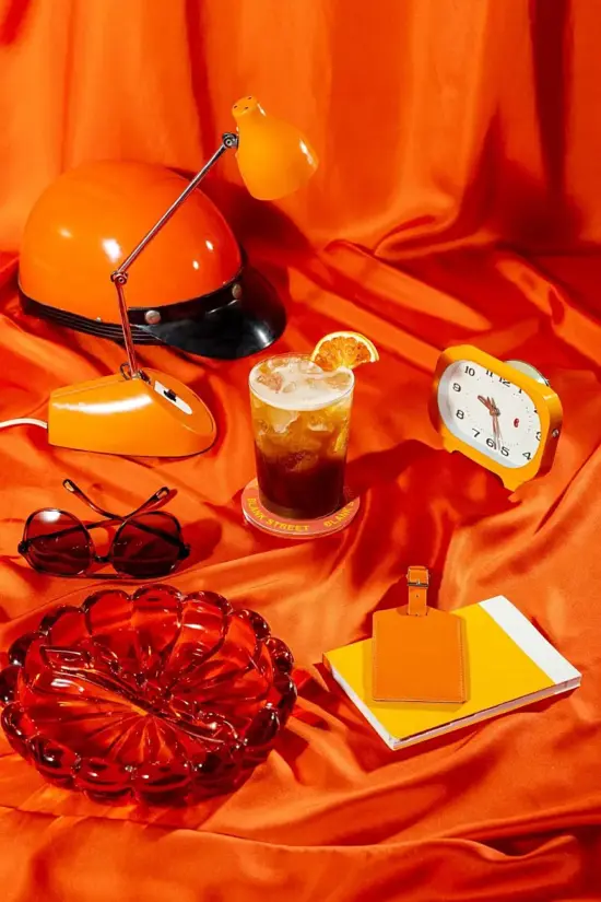 A cold coffee drink with an oranghe slice surrounded by random orange objects on a smooth satin orange cloth. 