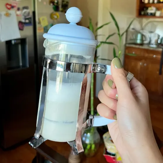 A hand with green nail polish holds up a small French press with frothed milk inside.