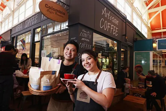 Two people hold up coffee mugs and bags outside the large market stall of Colombia Coffee Roaster, which has seating outside the structure and large windows looking in.