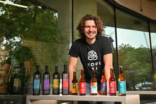 A man smiles in front of Kofi Ti's line of products, the bottles lined up in front of him on a table.