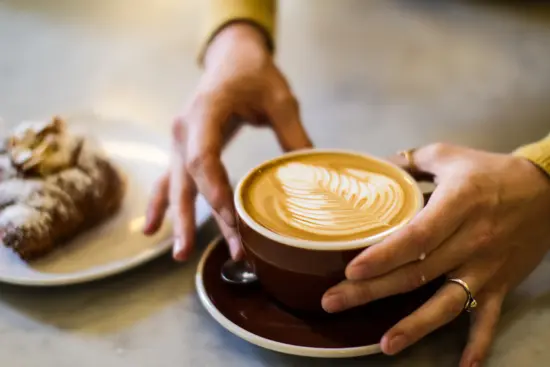 Two hands around a latte mug next to a pastry with powdered sugar.