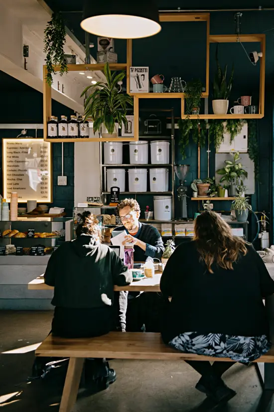 People sit at a table with benches inside the café. Buckets of coffee and the bar are visible behind them, while geometric shleves hold merchandise and plants.