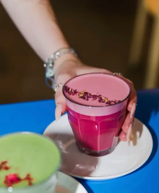 A hand sets down a beetroot latte in a glass cup next to a matcha on a blue table.