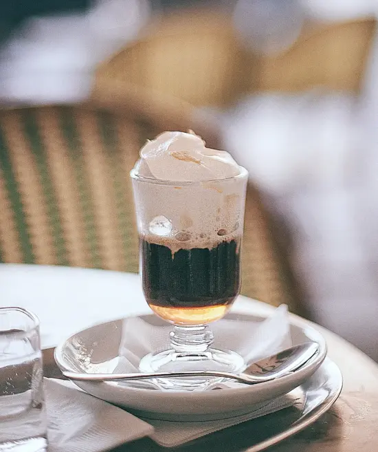 Irish coffee with a large heap of whipped cream on top, in a traditional Irish coffee glass with handle. It sits on a saucer with a spoon.