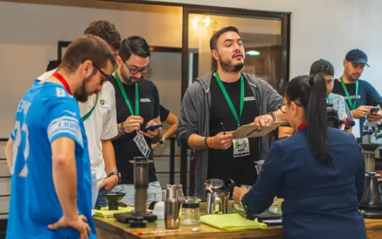 Judges hold clipboards. AeroPress and coffee paraphernalia are on the table.