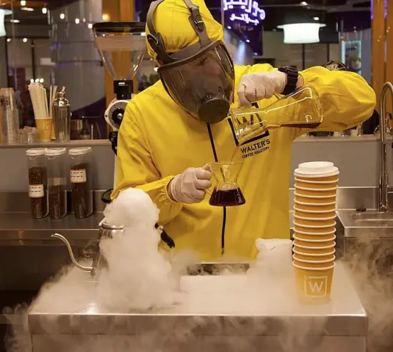 A barista in a yellow hazmat suit pours coffee into a beaker.