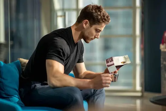 Drew Dennehy sits on a blue chair and looks at a coffee bag, holding a metal cup.