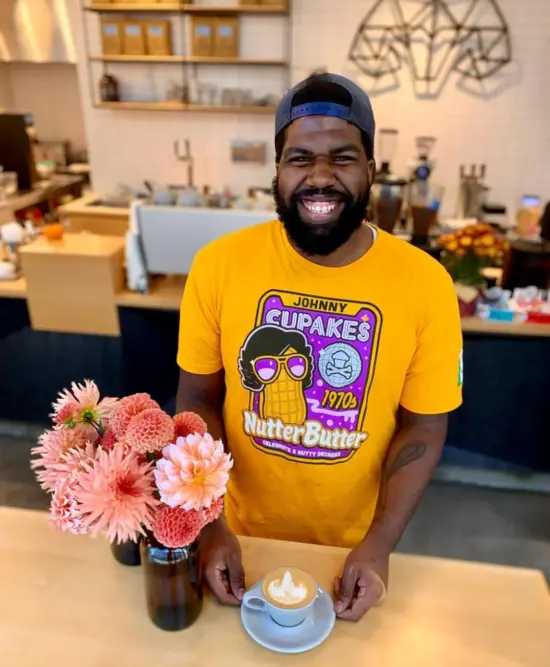 A person with a yellow t shirt next to a bouquet of pink flowers. He holds a latte in a mug on a saucer.