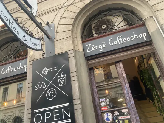 Zërgë Coffeeshop's storefront has an arched window above large doors, stickers on the door, and an "open" sign with portafilter, coffee, croissant, and bagel illustrations.