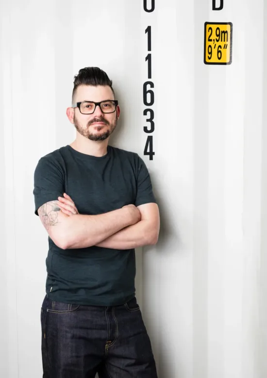 person woth glasses and arms folded leans against a shipping container.