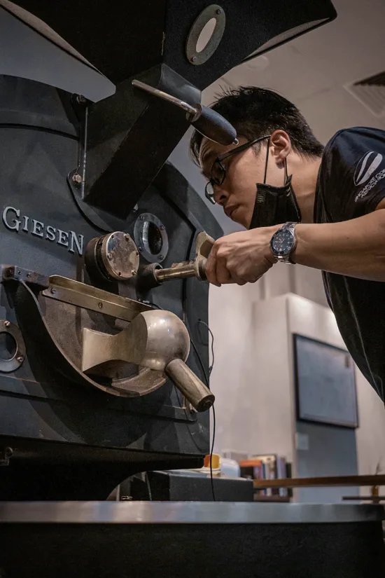 A roasting person checks beans from out of the roaster.