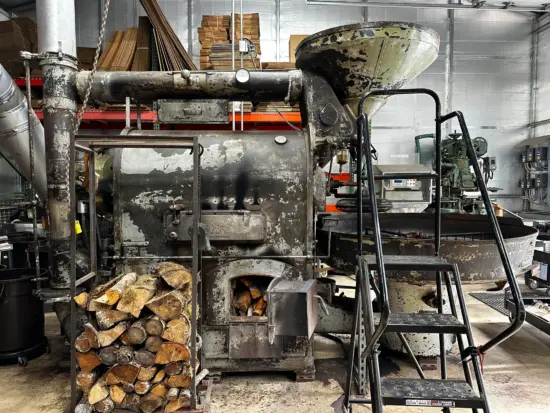 The huge metal roaster has an attached wood fire stove underneath, a large funnel up top to pour green beans into, and a huge round roasting  tray in the front.