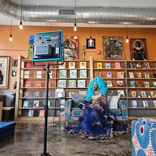 A drag queen in blue gown and wig sits with a book in Rofhiwa Book Cafe, with books lined along the wall behind them.