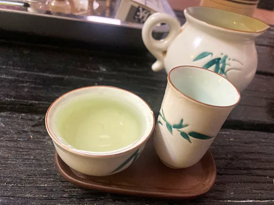 A cream-colored tea set, with a pot, short cup, and tall cup. Each is decorated with painted green leaves of bamboo.
