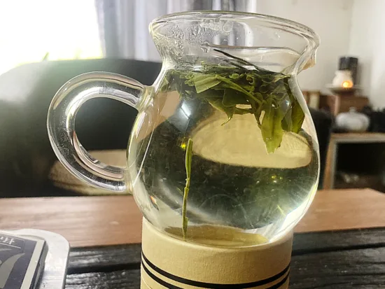 A clear glass teapot with green tea leaves.