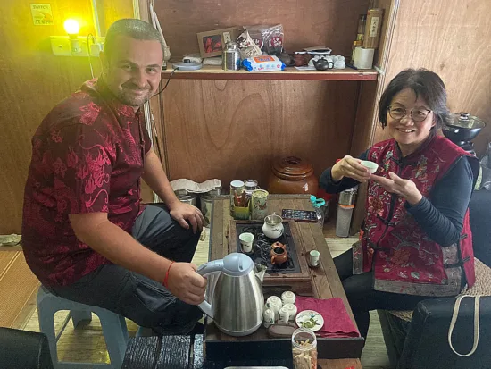 A student at the tea ceremony wears a red shirt. The tea master wears a red vest with embroidery and holds up a tiny teacup.