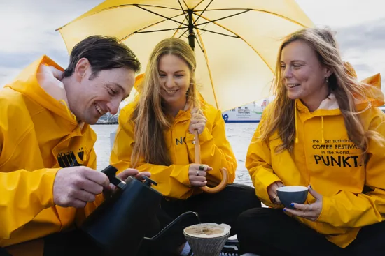 Steve and Anna W. and Anna J. sit together under a yellow umbrella, wearing matching yellow rain jackets, while Steve brews coffee with a pourover.