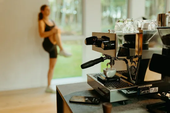 A woman works out in the background behind a home coffee machine pulling a shot.