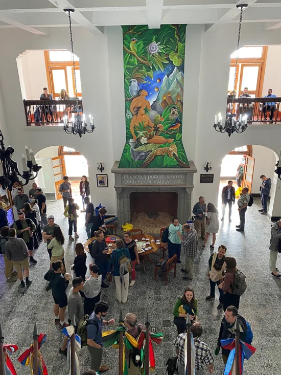 A large hall with a painting of a man and woman with jungle animals above the large fireplace and people standing around below it.