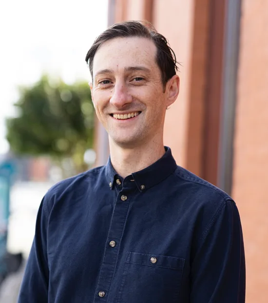 Steve Moloney stands outside a peach colored building, wears a dark blue button up shirt, and smiles for the camera.