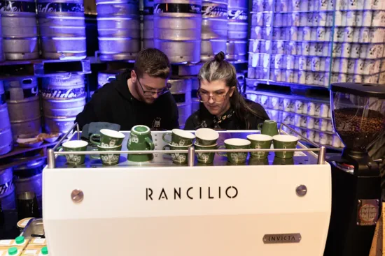 Two baristas behind a white Rancilio espresso machine at an Atlanta event. Giant kegs are stacked up behind them.