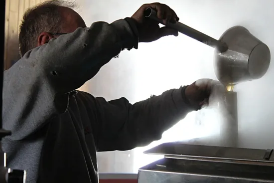 A man pours maple syrup from a large ladle into a metal tube for processing as steam rises up.