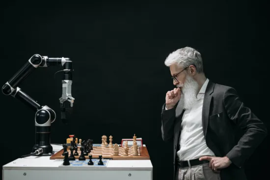 A bearded man ponders a chess move while a robot with arm stands on the table across from him, next to a chessboard.