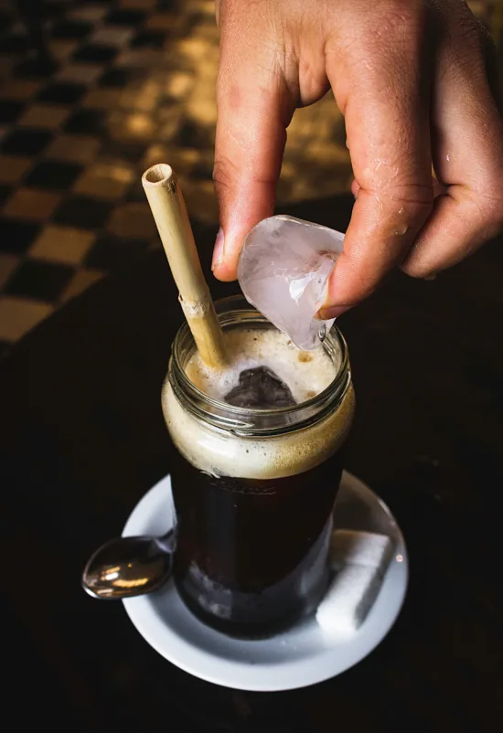 A glass jar with iced coffee and a hand placing an ice cube in the glass. A saucer holds sugar cubes and spoon. A can straw sits in the glass jar.