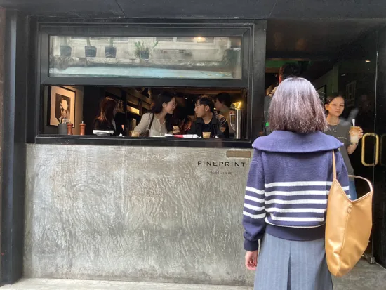 The exterior of Fineprint is concrete with a long rectangular window. A customer exits while a woman in purple sweater waits to enter.