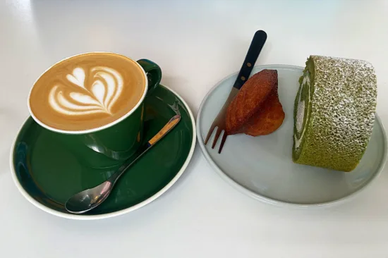 A green mug with a cortado, and a small plate with a green matcha cake roll and madeleine cookie.