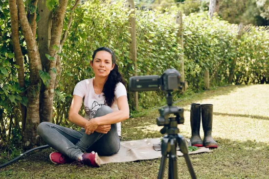 A woman sits on the ground speaking into a camera on a tripod.