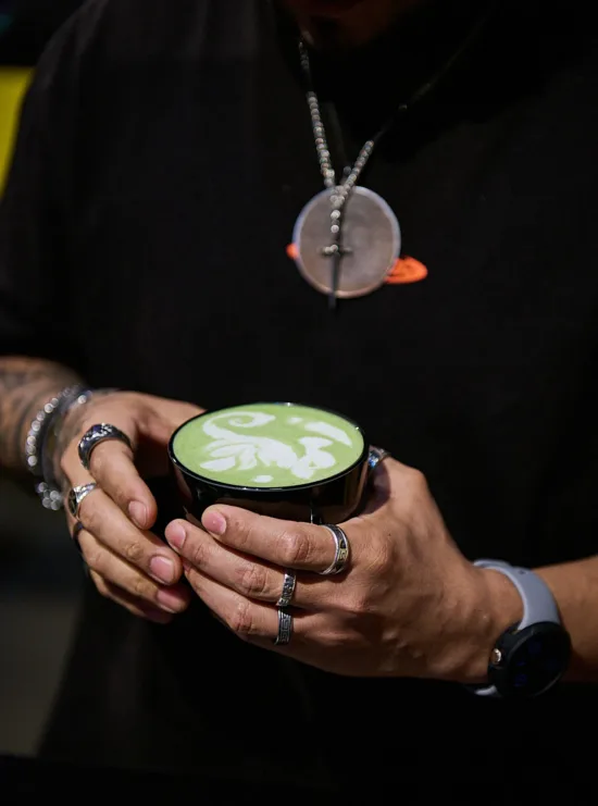 Two hands covered in silver rings hold a black mug with a seahorse design in a green matcha drink.