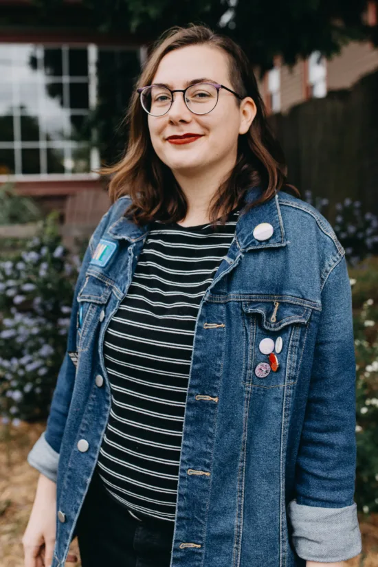 Brit stands outside with blue flowers behind, wearing a denim jacket and glasses.