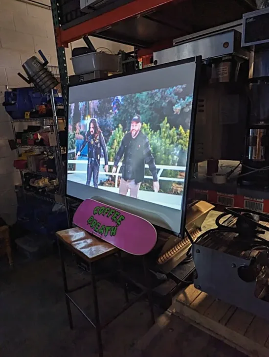 On a screen, two people stand with arms out. Below the screen is a skate deck in pink with "Coffee Breath" written on it in green slime font.