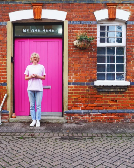 Em stands in front of a brick building with a coffee cup in hand. The door behind Em is hot pink and the words WE ARE HERE are printed on a small window above it.