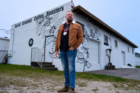 Jason stands outside a white building with an outline of a coffee plant painted on the corner, and the words Social Grounds Coffee.