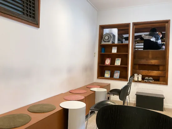 A bright room with low wooden bench with small white tables and black chairs, and built-in bookshelves.