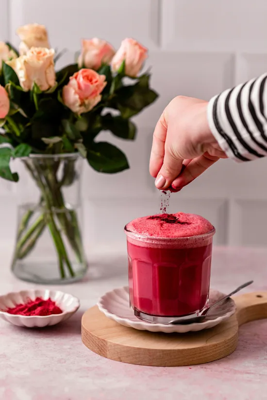 A dark pink drink, with a hand sprinkling beetroot powder on top, and orange and pink roses in a vase in the background.