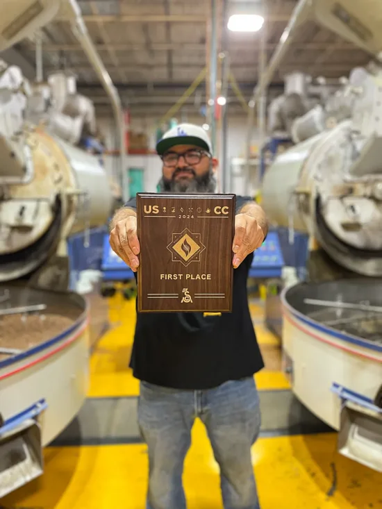 Eduardo standing between two roasting machines, with his USCC First Place plaque.