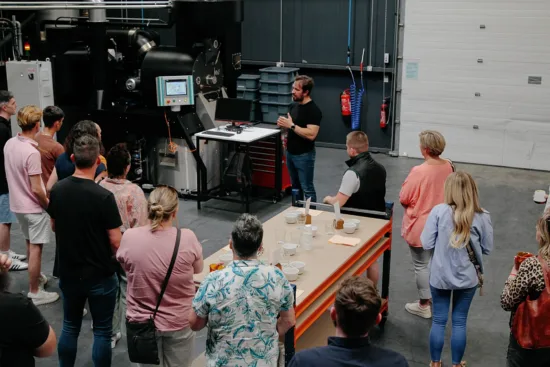 Nikola speaks to a group of people standing in a warehouse. A tasting table and roaster are inside the room.