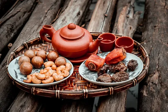 A red teapot and teacups with dried dates and other snacks on a woven tray.